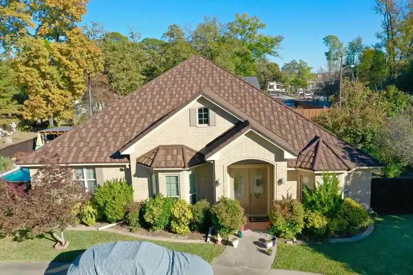 A brick home with a brown stone coated steel roof, autumn trees and a covered car in the drive