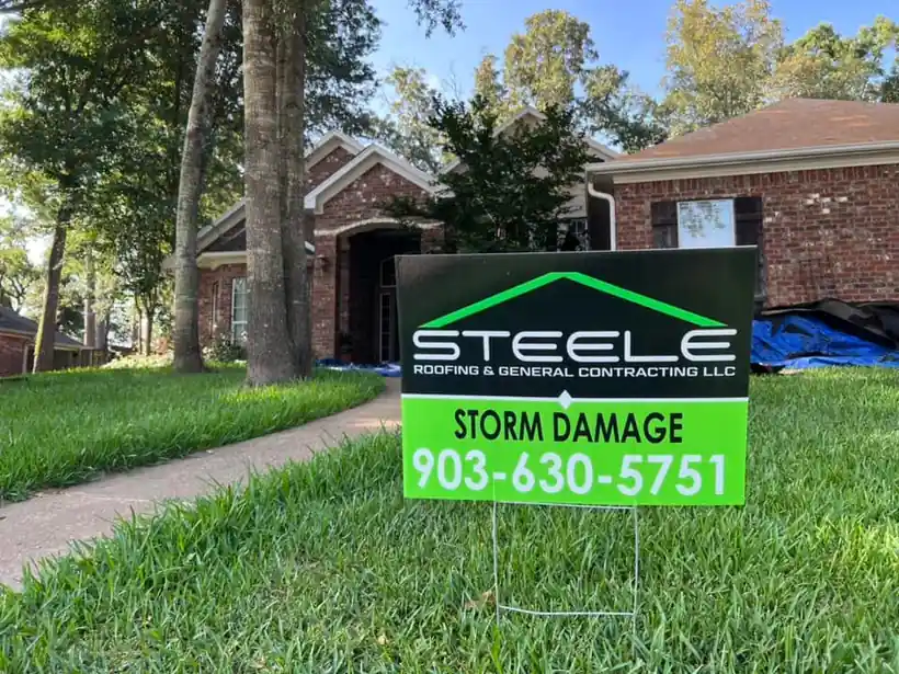A Steele Roofing storm damage yard sign on a lawn, a tarp on the house behind it