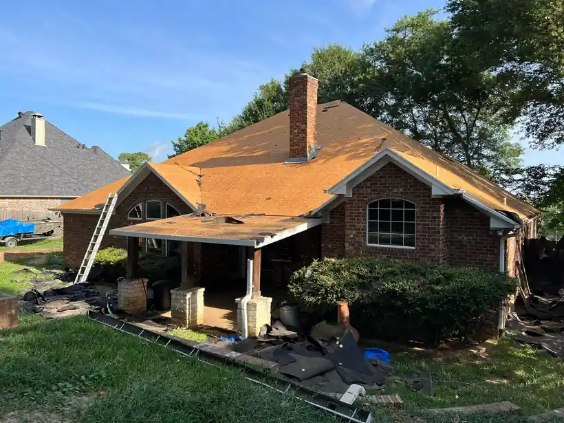 A brick home stripped down to bare decking mid tear-off, old shingles piled on the lawn